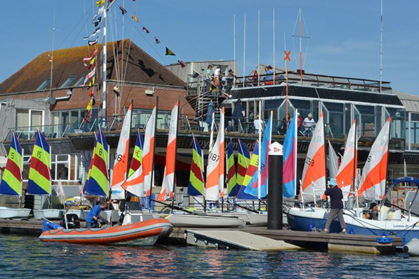 A group of boats with colourful sails at a sailing regatta outside a club house