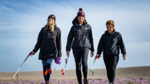 Three women are walking along a beach with litter pickers in their hands