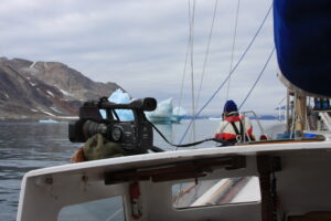 A cameraman is filming from the side of a boat. In the distance are icebergs. 